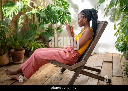 Giggling African American woman using phone. Happy girl relaxing at indoor home urban jungle. Stock Photo