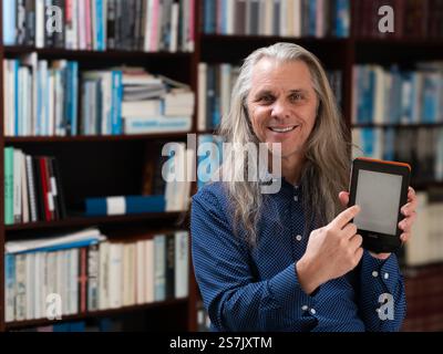 With a cheerful smile, a long-haired man proudly gestures toward his e-book, surrounded by a backdrop of books on a cozy shelf. Stock Photo