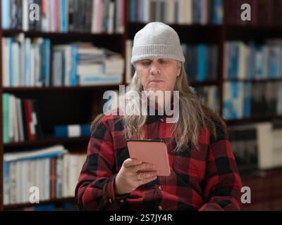 Immersed in a digital world, a long-haired man in a red plaid jacket and beanie sits by a bookshelf, absorbed in his e-book. Stock Photo