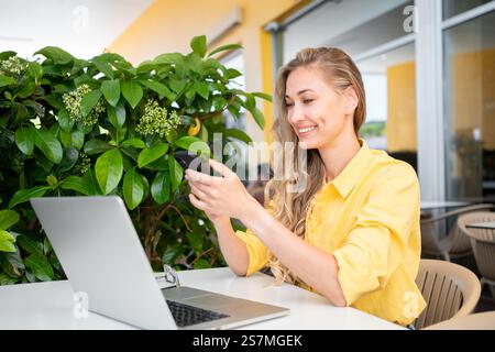 Happy freelancer woman using smartphone while sitting at restaurant. Beautiful female professional is with laptop on table during daytime outdoors Stock Photo