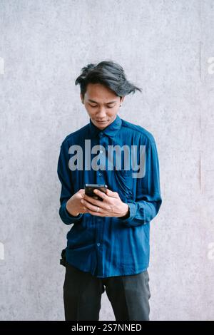 Young man focused on texting a message on his smartphone, standing against a wall. Stock Photo