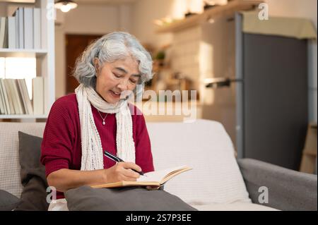 A happy, relaxed Asian retired woman enjoys writing in her diary while sitting on a sofa in the living room. retirement life, healthy living, wellbein Stock Photo