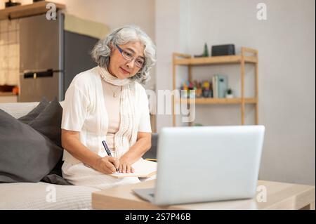 A positive, smiling Asian senior woman focuses on studying an online lesson through her laptop at home, writing notes from the lecture while sitting o Stock Photo