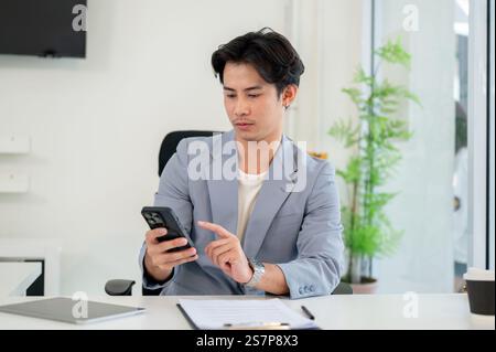 A handsome, smart Asian businessman or CEO in a business suit sits at his desk, focused on checking messages on his smartphone. people and wireless te Stock Photo