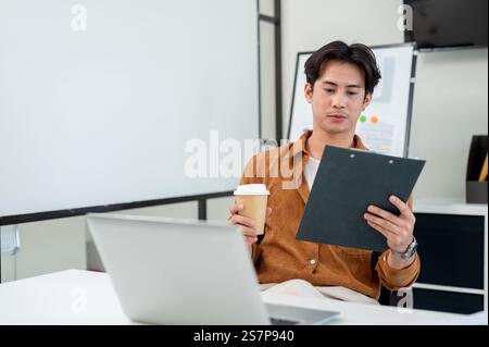A smart, handsome Asian male office worker focuses on reading a document at his desk in the office. corporate job, marketing agency, businessman Stock Photo