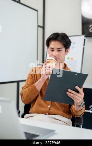 A smart, handsome Asian male office worker focuses on reading a document at his desk in the office. corporate job, marketing agency, businessman Stock Photo