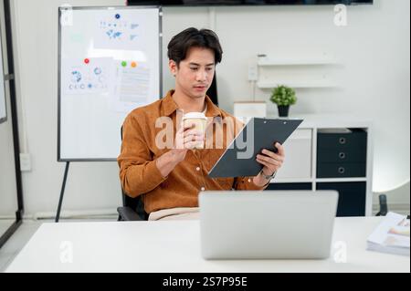 A smart, handsome Asian male office worker focuses on reading a document at his desk in the office. corporate job, marketing agency, businessman Stock Photo