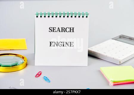 A notebook with the words search engine sits on a desk surrounded by office supplies and a calculator, illustrating technology's role in business. Stock Photo