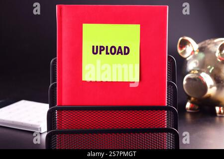 A bright red notebook sits on a black desk, featuring a neon yellow sticky note that reads upload. A golden piggy bank and a white notepad are also pr Stock Photo