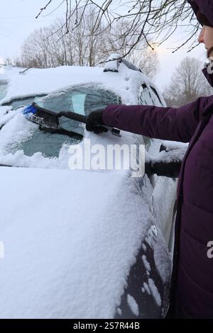 Side view of a young woman in warm clothes cleaning snow from a car with a special brush vertical orientation Stock Photo