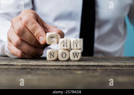 A hand in business attire places wooden blocks spelling TRY NOW on a rustic surface, symbolizing initiative and seizing opportunities. Stock Photo