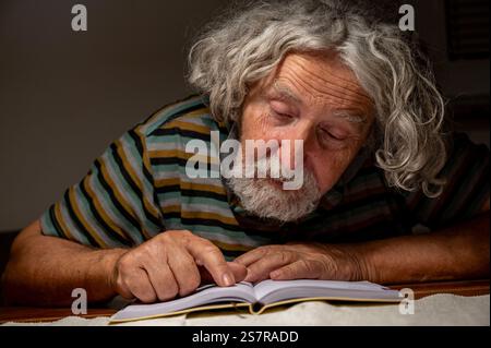 An older man with curly gray hair and a striped shirt leans over a book, pointing at the text, engrossed in reading under soft indoor lighting. Stock Photo