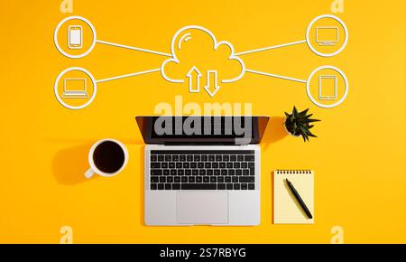 Cloud computing diagram showing data exchange between laptop, smartphone, tablet and computer on a yellow background, with coffee, notepad and pen Stock Photo