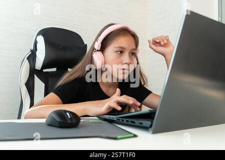 baby girl wearing headphone to listen music or using white keyboard laptop and study online on wood table desk in living room at home. Education learn Stock Photo