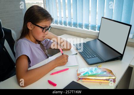 Primary school Student child writing in book using laptop. Distance learning online education. girl studying at home with digital tablet computer and Stock Photo