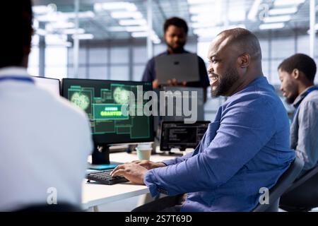 Cheerful engineer in data center getting access granted notification on PC screen, managing to access server files after they were locked by hacker using ransomware, receiving help from colleagues Stock Photo