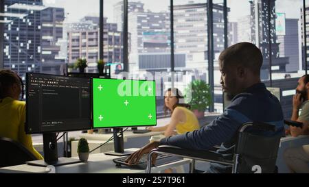Person with disability testing and deploying programs and systems on green screen PC. Software technician in wheelchair checking code on mockup PC, sitting at desk in office, camera B Stock Photo