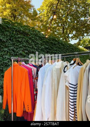 Colorful clothes hanging on a rack in a garden. Wardrobe items on hangers for sale at a local flea market - trendy second hand clothes for sale. Stock Photo