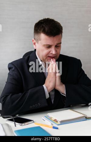 A businessman prays sitting at his desk in the office, focused on completing a difficult task, solving a problem. Stock Photo