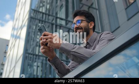 Smiling Arabian Indian man businessman entrepreneur manager use mobile phone application in city office terrace bottom view male business owner Stock Photo