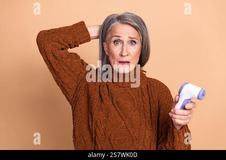 Elderly woman with surprised expression holding thermometer, against beige background Stock Photo