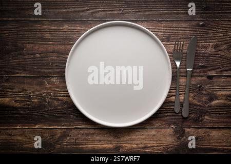 Top view of empty ceramic white plate, fork and table knife on wooden table Stock Photo