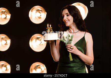 A smiling woman in a luxurious green dress presents a small birthday cake with '35' candles. She's holding white flowers, creating an elegant and fest Stock Photo
