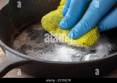 Person cleaning a rusty old cast iron frying pan using bicarbonate of soda and a scouring pad. On a wood board background. Stock Photo