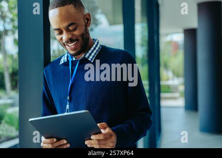 A young African businessman is seen using a tablet with a smile, standing in an office corridor. His confident demeanor suggests a productive work env Stock Photo