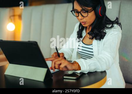 A woman wearing headphones engages with a tablet using a stylus, illustrating concentration and modern work in a cozy coworking environment. Stock Photo