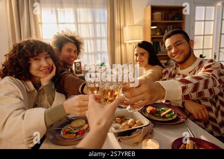 Unrecognizable hand clinking glass full of champagne with beloved friends who sitting in front of him at festive table with homemade food and decorations Stock Photo
