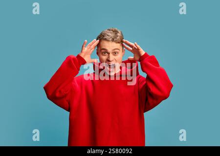 Portrait of young attractive man with surprised expression, open mouth and putting hands on head against blue studio background. Stock Photo