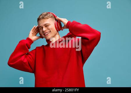 Young handsome man, dressed casual, listening music in headphones, expressing relax and joy against blue studio background. Stock Photo