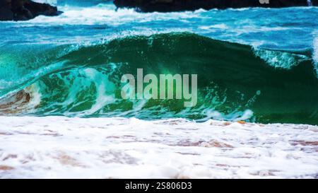 A powerful, emerald-green wave crashes onto the shore, creating a frothy white expanse of foam.  The wave's force is evident in the way it's breaking Stock Photo