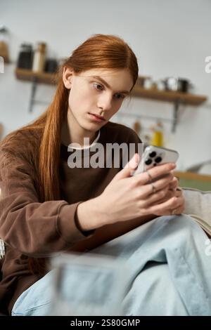 Stylish young man with long red hair relaxes on a couch while using his smartphone indoors. Stock Photo