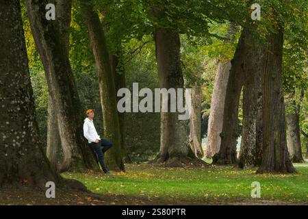 Young man leaning on an old tree in a scenic park with lush colors and enjoying the beauty of nature Stock Photo