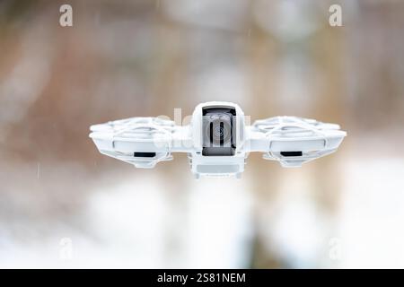 A white drone with a camera on it is flying in the air. The drone is capturing a view of the snow-covered ground below Stock Photo