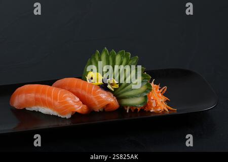 Two pieces of salmon nigiri sushi served on a black plate, garnished with a fan of cucumber slices, edible flowers, and shredded vegetables. Stock Photo