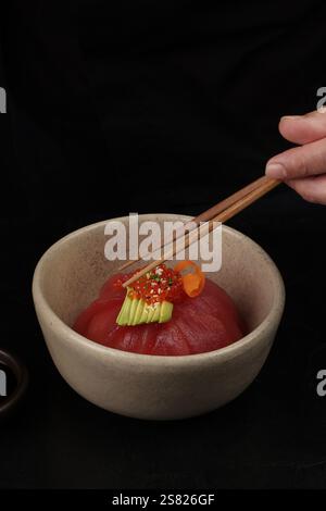 A delicate tuna dome topped with avocado slices, salmon roe, and carrot ribbons, served in a ceramic bowl, captured with a hand holding chopsticks for Stock Photo