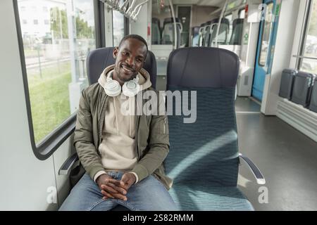 African American man sitting in the window seat on the moving train, using a smartphone and headphones to listen to music. Stock Photo
