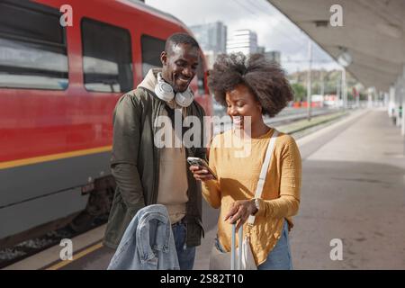 Smiling African American couple having fun using a smartphone while waiting for the train at the railway station platform. Stock Photo