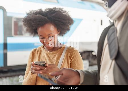 Smiling African American tourist couple with luggage using smartphone while waiting for a train on a railway station platform. Stock Photo