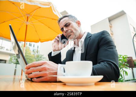 Determined Businessman Talking on a Call Conversation on His Cellphone While Engaged with His Laptop, Combining Productivity and Communication for Effective Work Performance in a Outdoor Cafe. High quality photo Stock Photo
