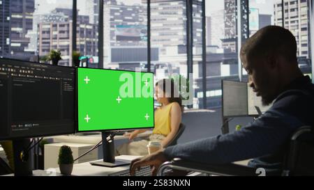 Man with disability in inclusive workplace working with machine learning tech on green screen computer. African american IT expert with paraplegia in wheelchair works with data on mockup PC, camera B Stock Photo