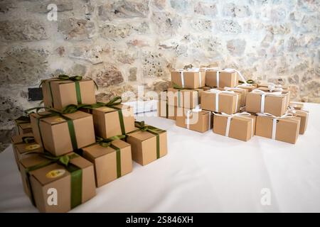 Two stacks of small brown gift boxes tied with green and white ribbons sit on a white tablecloth against a textured stone wall backdrop. Stock Photo