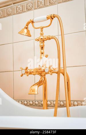 Gold colored bathtub faucet with white ceramic details and handheld shower, mounted on beige tiles with a floral decorative border. Stock Photo