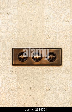 A brass triple toggle light switch panel with rounded toggles mounted on a wall of cream colored floral and vine patterned wallpaper. Stock Photo