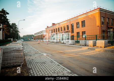 Partially paved road with construction materials near a modern two story building. Streetlights, traffic signs, and trees visible in the background. Stock Photo