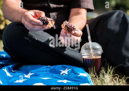 A person sits on a blue blanket with star patterns, holding a split chocolate pastry. A plastic cup with iced coffee rests nearby on the grass. Stock Photo