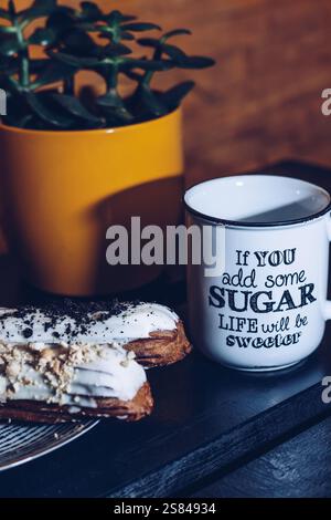 Close up of a white mug with black text, two eclairs with white icing, and a yellow pot holding a green succulent on a dark surface. Stock Photo